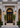 A grand black double front door with ornate ironwork and glass panels, set in a beige stone building entrance with two potted green trees on either side.