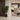 A tall green potted plant in a black geometric planter stands in a modern interior hallway beside a dark wood door, with a bright living room and dining area visible in the background.