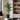 A tall artificial palm tree in a black geometric pot, placed in a modern living room corner next to a beige sofa and a built-in shelving unit.