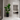 A large potted bird of paradise plant with broad green leaves stands in a black planter on a white marble floor, positioned in a modern hallway next to a dark wood door, with an open doorway revealing a living room in the background.