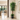 A tall artificial indoor tree with lush green leaves and a twisted trunk, styled in a dark gray geometric pot on a light tiled floor in a modern home hallway.