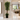 A tall artificial potted tree with lush green leaves and a textured brown trunk stands in a modern, upscale hallway. The black planter sits on a polished light beige marble floor, with a cozy living room visible in the background where two people are seated on a sofa.