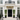 A grand white stucco mansion facade with a black double front door, white columns, a black wrought-iron balcony, and two tall potted green trees flanking the entrance.