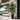 A tall artificial palm tree with lush green fronds in a black geometric planter, placed on a modern outdoor patio next to a concrete wall.