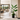 A large potted houseplant with broad green leaves and smaller trailing greenery in a black pot, placed in a modern interior hallway with a living room visible in the background.