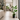 A tall green potted plant with large tropical leaves stands in a modern, bright living room with light wood floors, a stone pillar, and a staircase in the background.